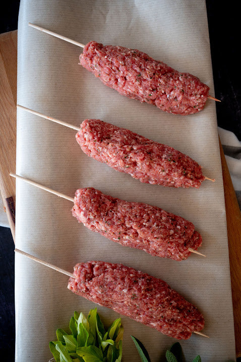A set of four uncooked lamb koftas skewered on wooden sticks, placed on a beige surface with herbs nearby.