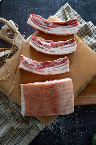 Raw belly pork cuts on paper and a wooden board, with a kitchen towel in the background.