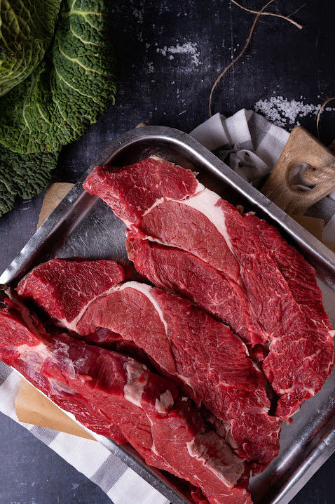 Raw braising steak cuts on a metal tray with a head of lettuce in the background.