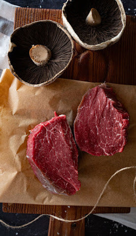 Two raw steaks and two mushrooms on a wooden cutting board with brown paper underneath.