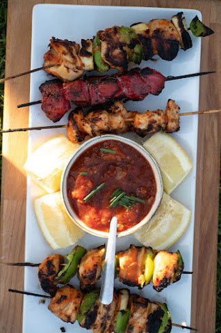 Platter of skewered grilled meats with a bowl of red sauce and lemon wedges on a wooden surface.
