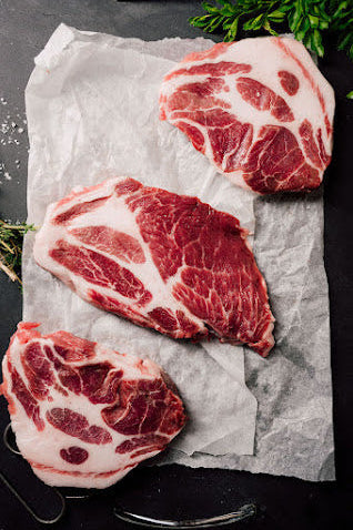 Three fresh prime rib steaks on a piece of paper, with herbs and spices around, on a dark background.