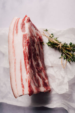A piece of Mangalitza belly pork with visible marbling, placed on a white surface, accompanied by a sprig of thyme.