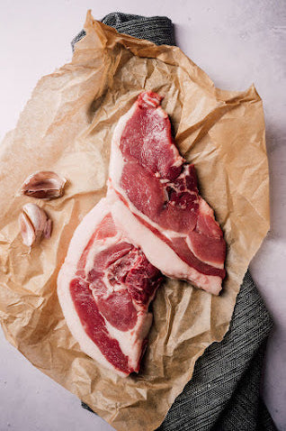 Two fresh Mangalitza leg steaks on a piece of brown paper, with a garlic clove and a twist of pepper in the background.