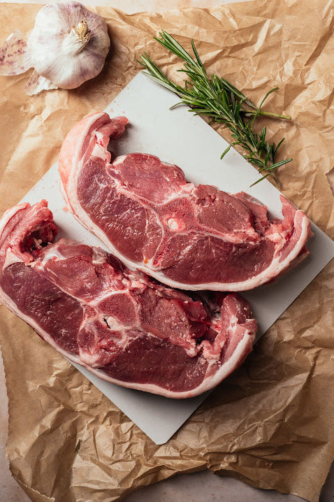 Two fresh double lamb chops on a white plate with a sprig of rosemary and a garlic clove, presented on a brown paper background.