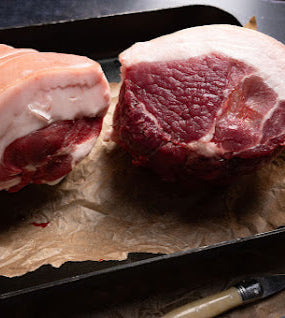 A rolled leg joint of pork, still with the skin on, tied at one end, placed on a baking tray with a piece of brown paper underneath. The joint is pink in color with visible fat marbling.