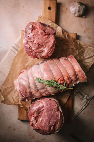 A tied rolled lamb shoulder joint on paper and aa wooden boardwith rosemary sprigs and garlic clove in the background.