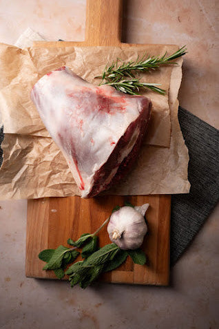 A fresh lamb shank on a piece of brown paper, accompanied by a sprig of rosemary and a garlic clove, placed on a wooden board.