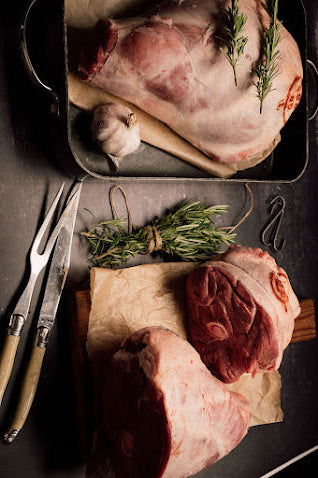 RAW LEGS OF LAMB ON DARK SURFACE WITH ROSEMARY SPRIGS AND CARVING SET