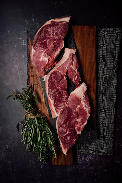 Raw lamb steaks on a wooden cutting board with a sprig of rosemary beside them, all placed on a dark, textured surface.