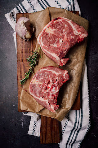 Two raw lamb neck cuts on butchers paper and a wooden board, accompanied by a sprig of rosemary and a clove of garlic.