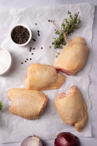 Several boneless, skinless free-range chicken thighs on a white surface with salt, pepper, onion, and herbs.