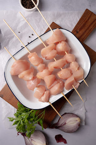 Raw chicken skewers on a white tin dish with skewers made of wooden sticks, surrounded by garnishes including green parsley and red onion.