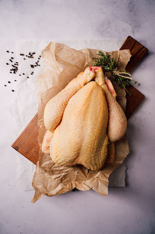A whole free-range chicken on a wooden board, covered with a paper, with herbs and spices scattered around, ready for cooking.