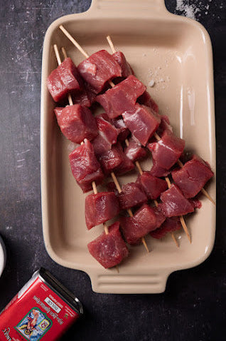 Raw minted lamb skewers on a beige tray, with a tin of paprika and various spices visible in the background.