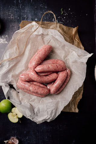 A pack of six fresh Mangalitza pork and apple sausages on a piece of wrapping paper, with apple pieces and greenery in the background.
