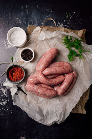 A set of uncooked Mangalitza Cumberland sausages on a paper, seasoned with herbs and spices, including a small bowl of red pepper, a dish of salt, and some fresh green parsley.