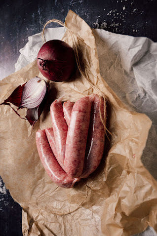 Raw Mangalitza caramelised onion sausages on a piece of brown paper, accompanied by a shallot and an onion.